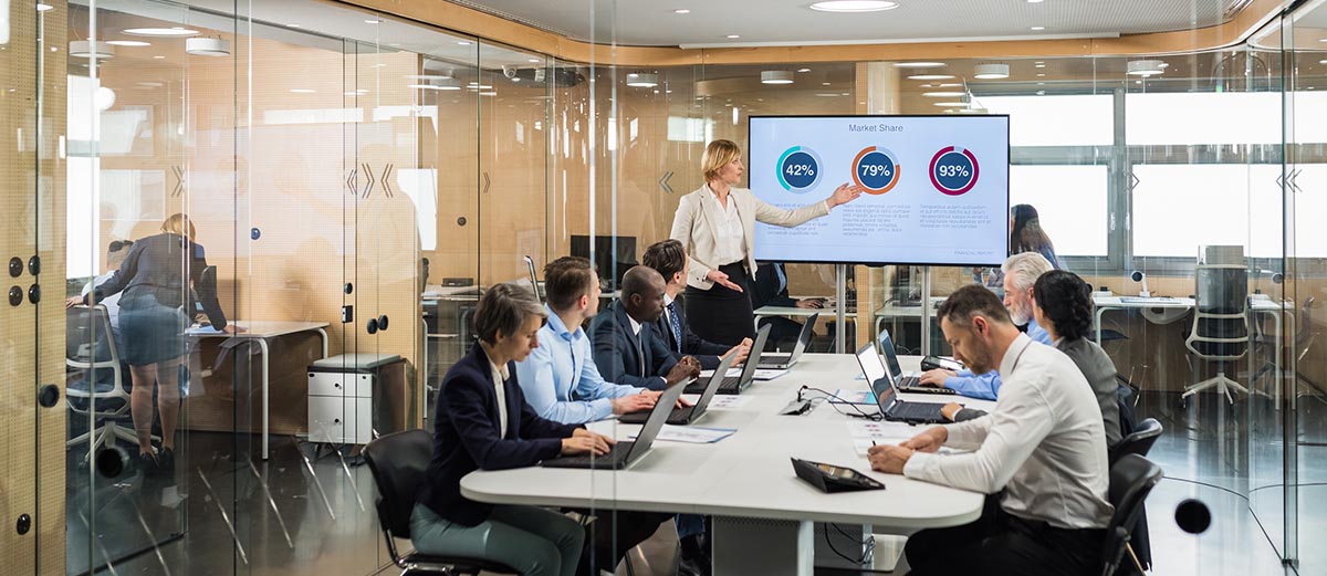 Eight people in a corporate board room, seven of them are sitting around the table watching and listening to the eighth person give a presentation on a large screen that is displaying numbers. 