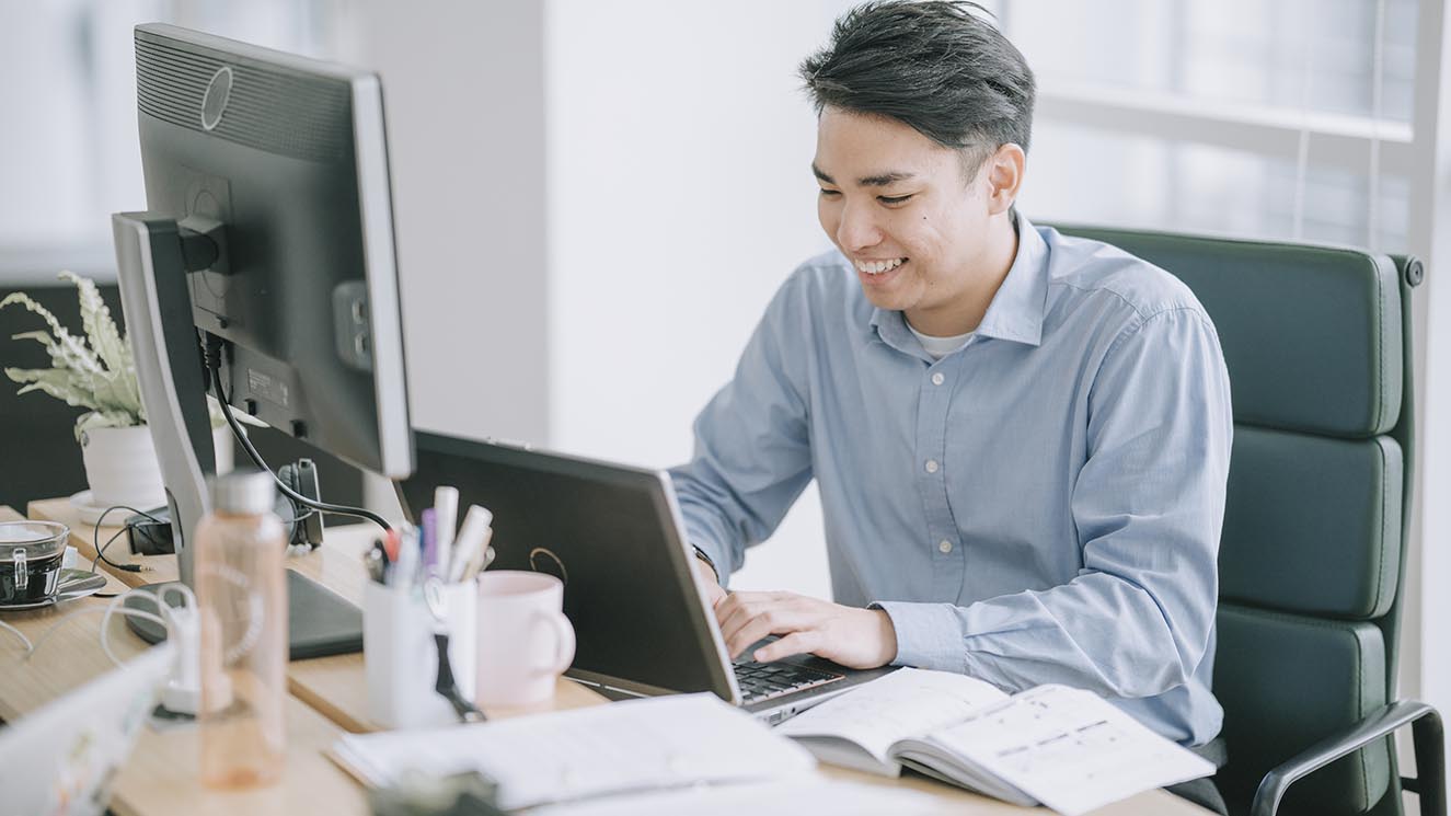 Young man in professional attire working at a desk where he has a laptop and a large computer monitor as he is working on escheatment tasks.