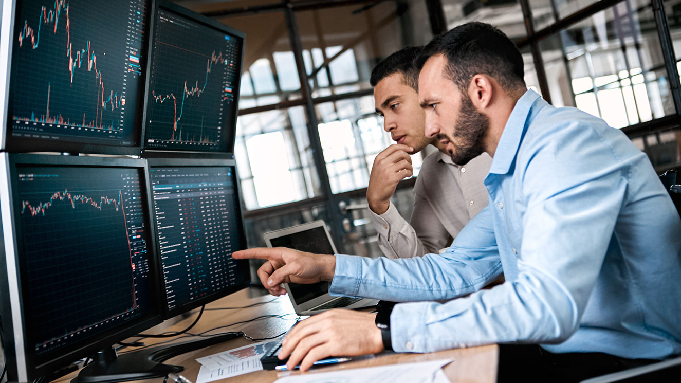 Two men at a desk looking at four computer monitors that are showing line graphs and charts.