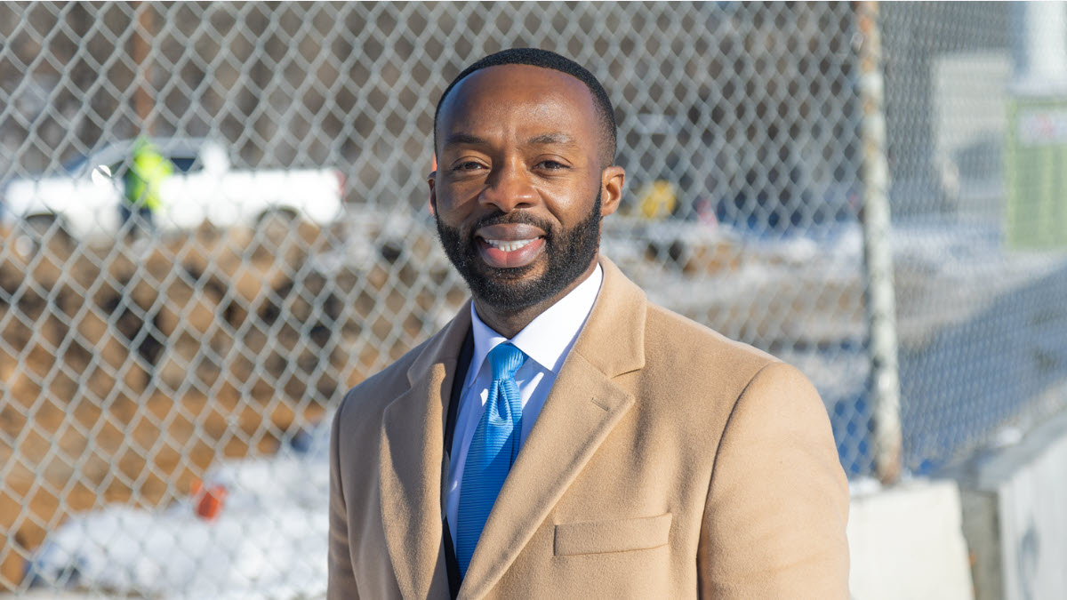 Professionally dressed man wearing a shirt, tie and overcoat standing in front of a construction site.