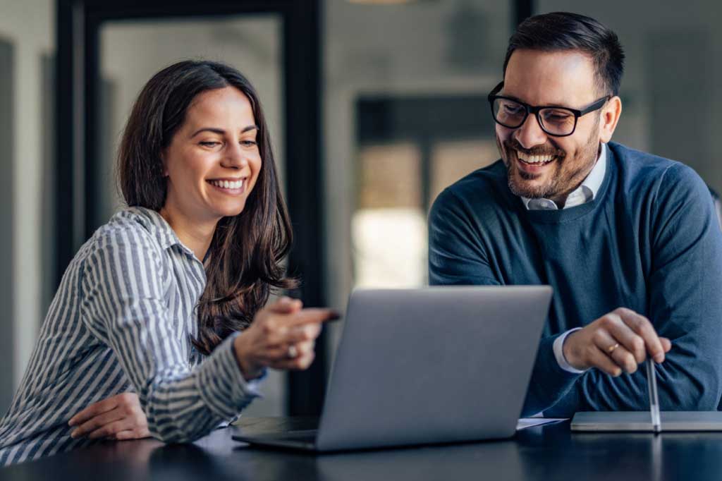 A man and woman sitting next to each other at a desk in an office and looking at a laptop.