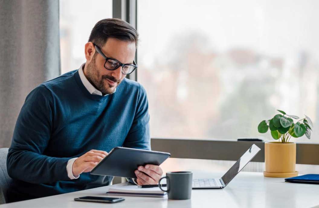 Man wearing glasses sitting at a desk in front of a laptop and looking at his tablet device at the same time.