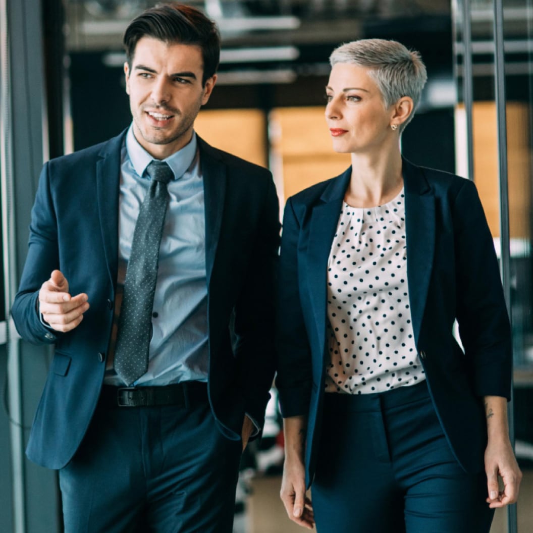 Male and female coworkers dressed in business suits and walking next to each other down a hallway in an office building.