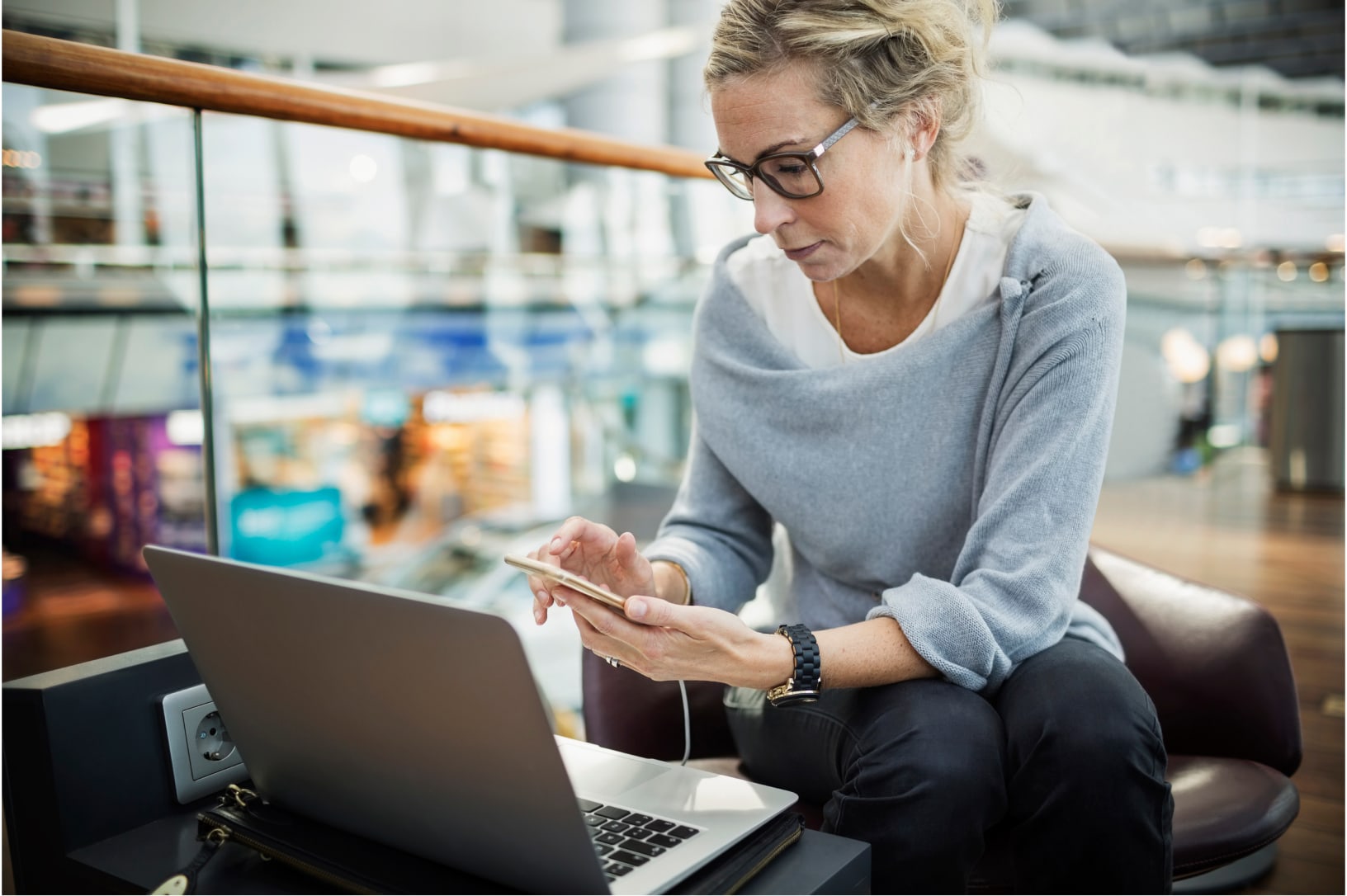 Woman wearing glasses sitting in a lounge chair with her laptop and cell phone, using her devices for payments.