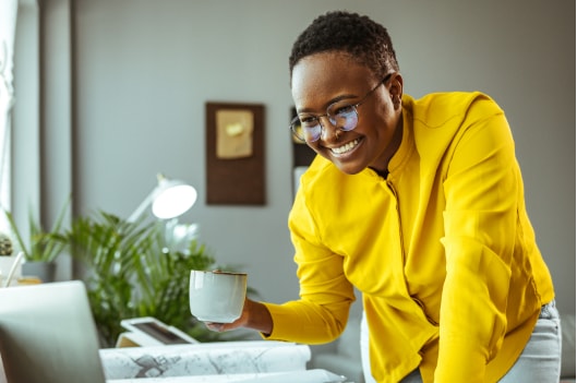 A young, brightly-dressed Black woman smiles at her laptop screen.