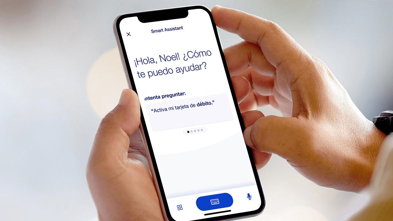 Pantalla de un teléfono celular que muestra la Aplicación Móvil de U.S. Bank en dirección a la cámara (solo se muestran las manos de la persona que lo sostiene). La pantalla dice: ‘¡Hola, Noel! ¿Como te puedo ayudar?’ y presenta un tema de conversación a continuación, también en español.