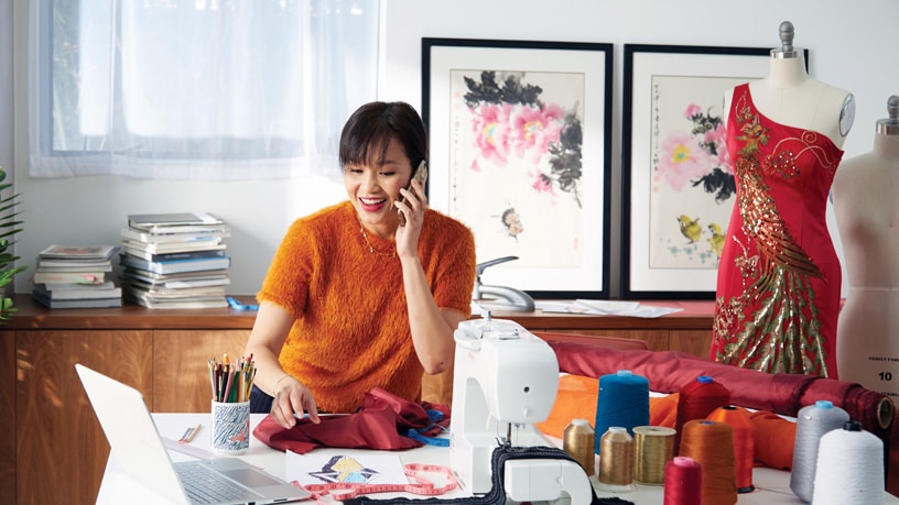 A young Chinese woman works on making a red dress accented with gold while talking on her phone.