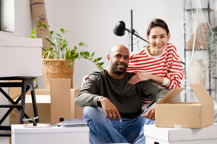 A man and woman are sitting and smiling in their living room while unpacking moving boxes.