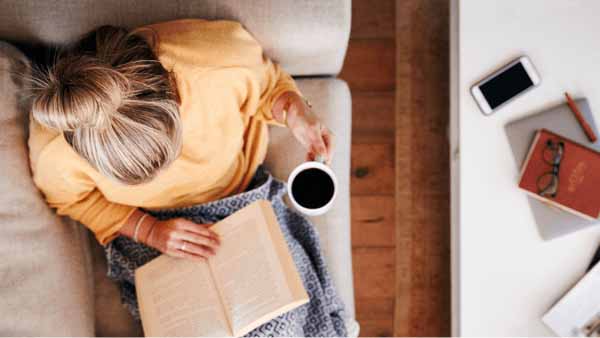 A woman relaxes on a couch, reading a book that compares FHA and conventional loans