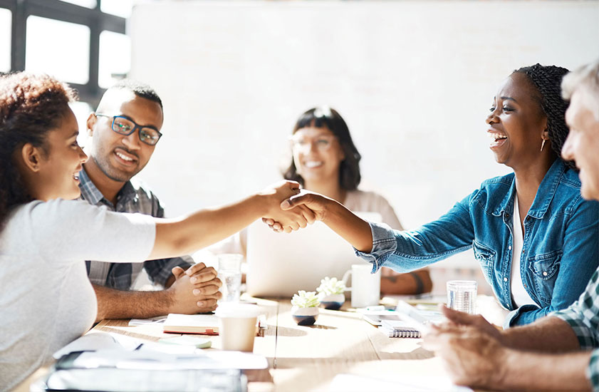 Five people sitting around a table and talking, with two of them reaching across the table to shake hands.