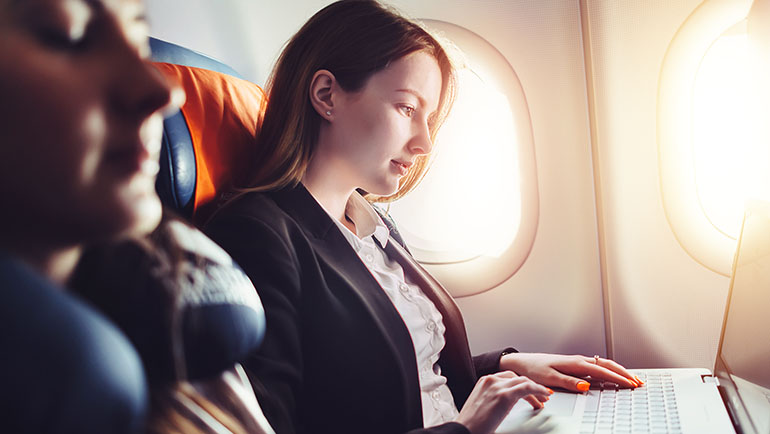 Woman working on laptop while sitting in a window seat on an airplane.