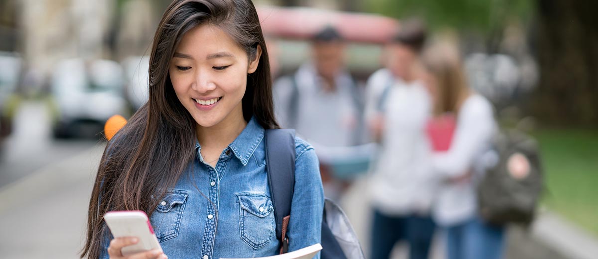 College-aged woman wearing a backpack and looking at her phone while walking on campus.