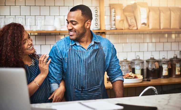 Man and woman in a large kitchen wearing aprons and smiling at each other.