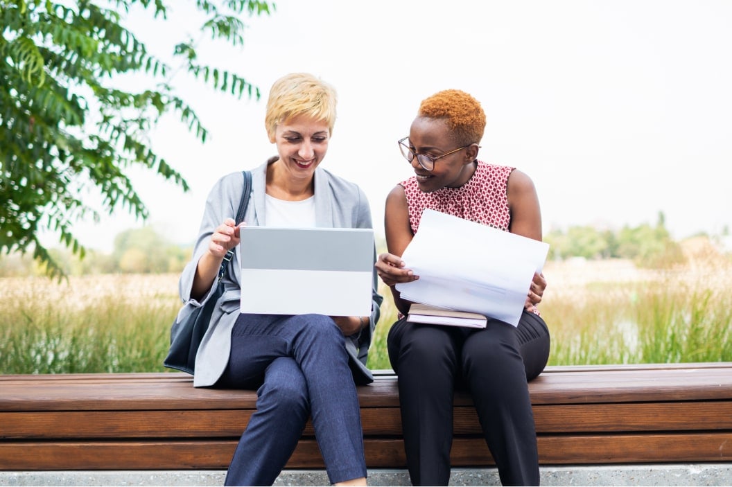 Two women sitting on a bench next to each other. One is holding paperwork while the other has a tablet open and they are doing business together.