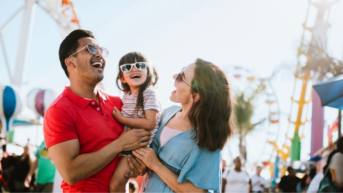 A Hispanic family including mom, dad and little girl smile and laugh together at a carnival.