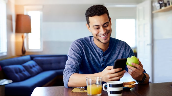 A young Hispanic man is viewing his phone, smiling while eating breakfast.