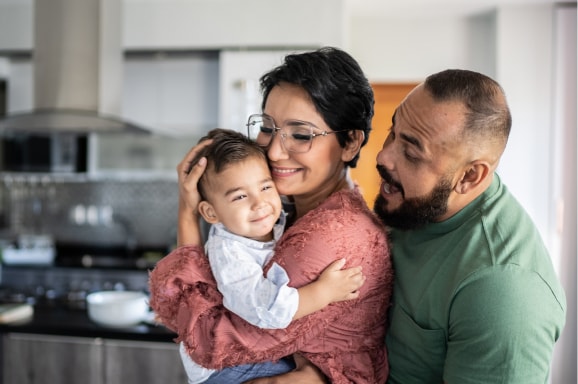 A family including mom, dad and baby snuggle together in their kitchen.