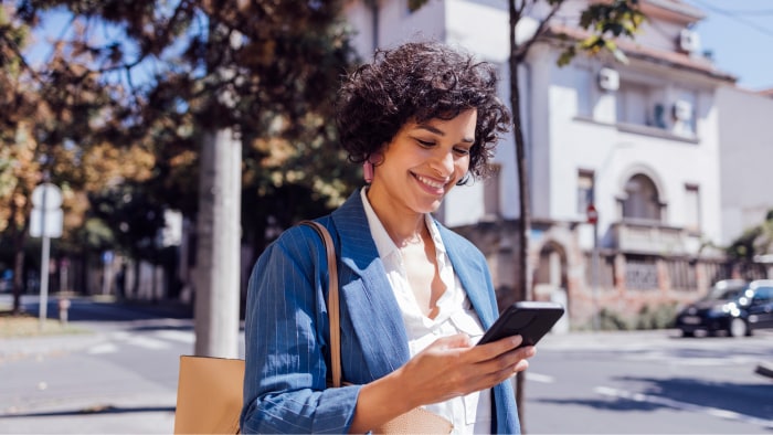 A.woman with short, dark hair wearing a blue jacket is standing on a street corner, looking down at her phone, smiling.